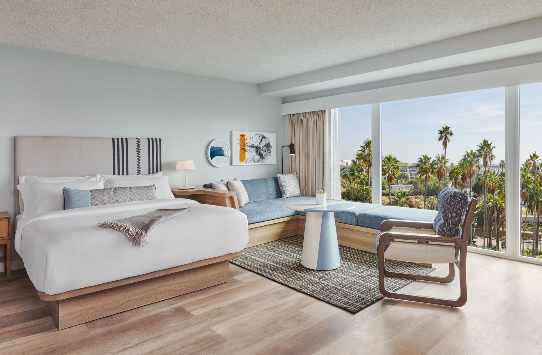 Hotel bedroom with a king bed, seating area, and large windows showing palm trees outside.