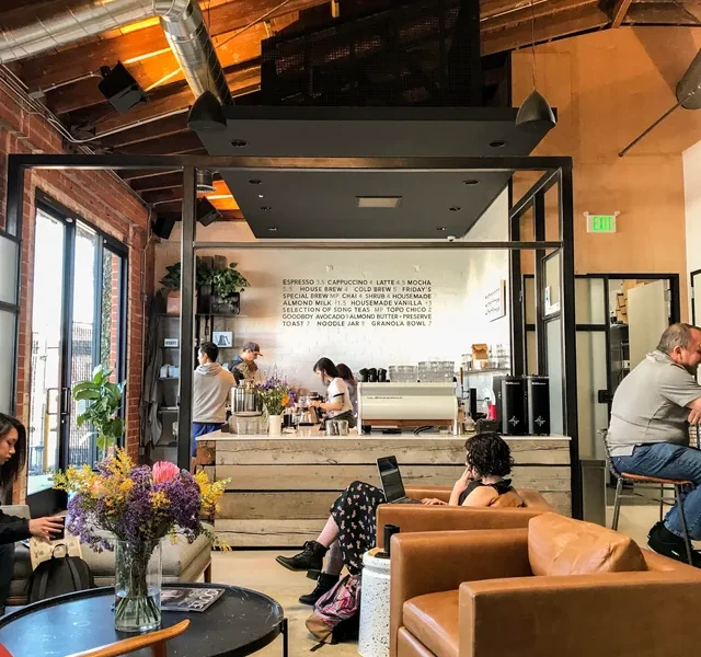Cozy industrial cafe with exposed beams and a service counter; patrons sit on leather and red chairs while a barista works behind the counter.