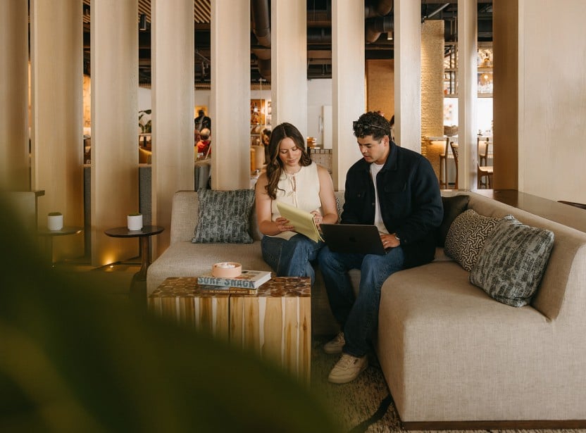 Two people sit on a beige sofa in a bright, modern lounge, looking at a yellow folder and a laptop together.
