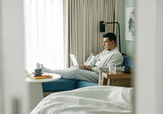 Man in light gray loungewear sitting on a blue sofa, typing on a laptop by a large window with curtains.
