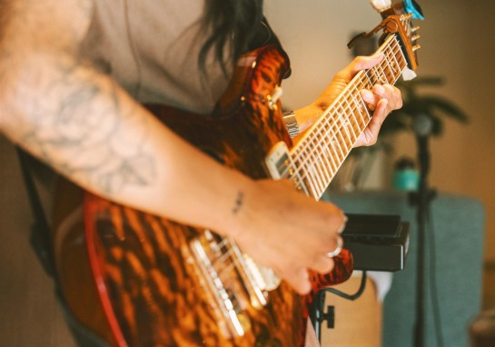 Close-up of a person playing an electric guitar, focusing on fingers pressing strings along the neck with a blurred background.