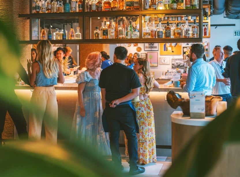 People gathered at a bar counter, conversing, with shelves full of assorted liquor bottles above them.
