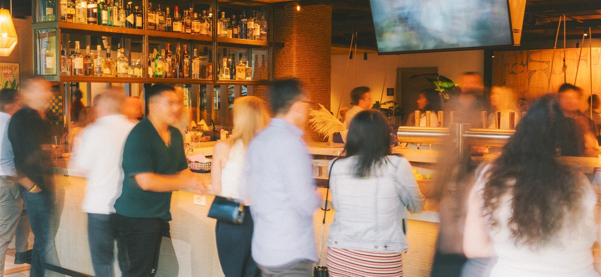 Busy bar interior with liquor shelves behind the counter, warm pendant lights, and patrons waiting at the bar.
