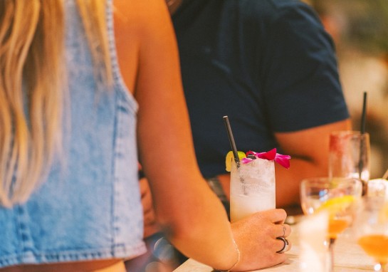 Close-up of a hand holding a frosty cocktail with a lime wedge and pink flower garnish, at a bar with blurred people in the background.