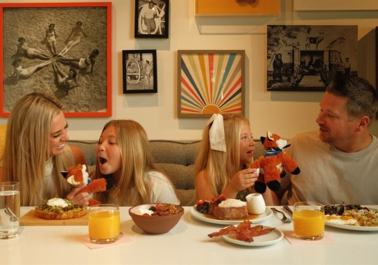Family of four at a dining table with food and orange juice, two girls holding plush foxes while a woman and man smile nearby in a cozy dining room with wall art.