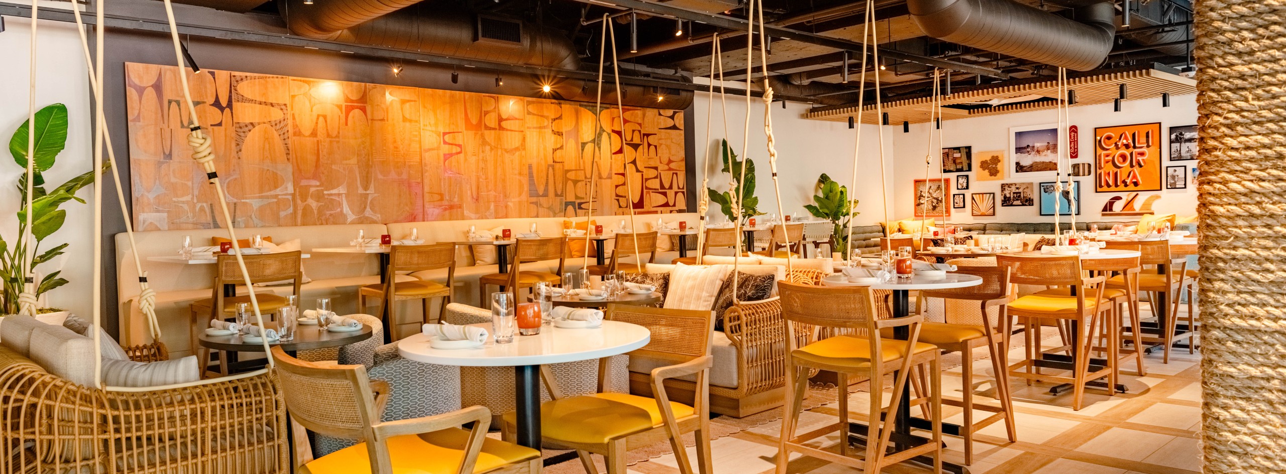 Restaurant interior with rope-swing seating, woven chairs, round tables, warm lighting, and tropical plants along the walls/left side.