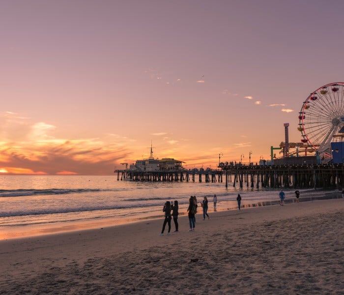 Sunset over the ocean with a lit Ferris wheel on a pier to the right and people walking along the shore.