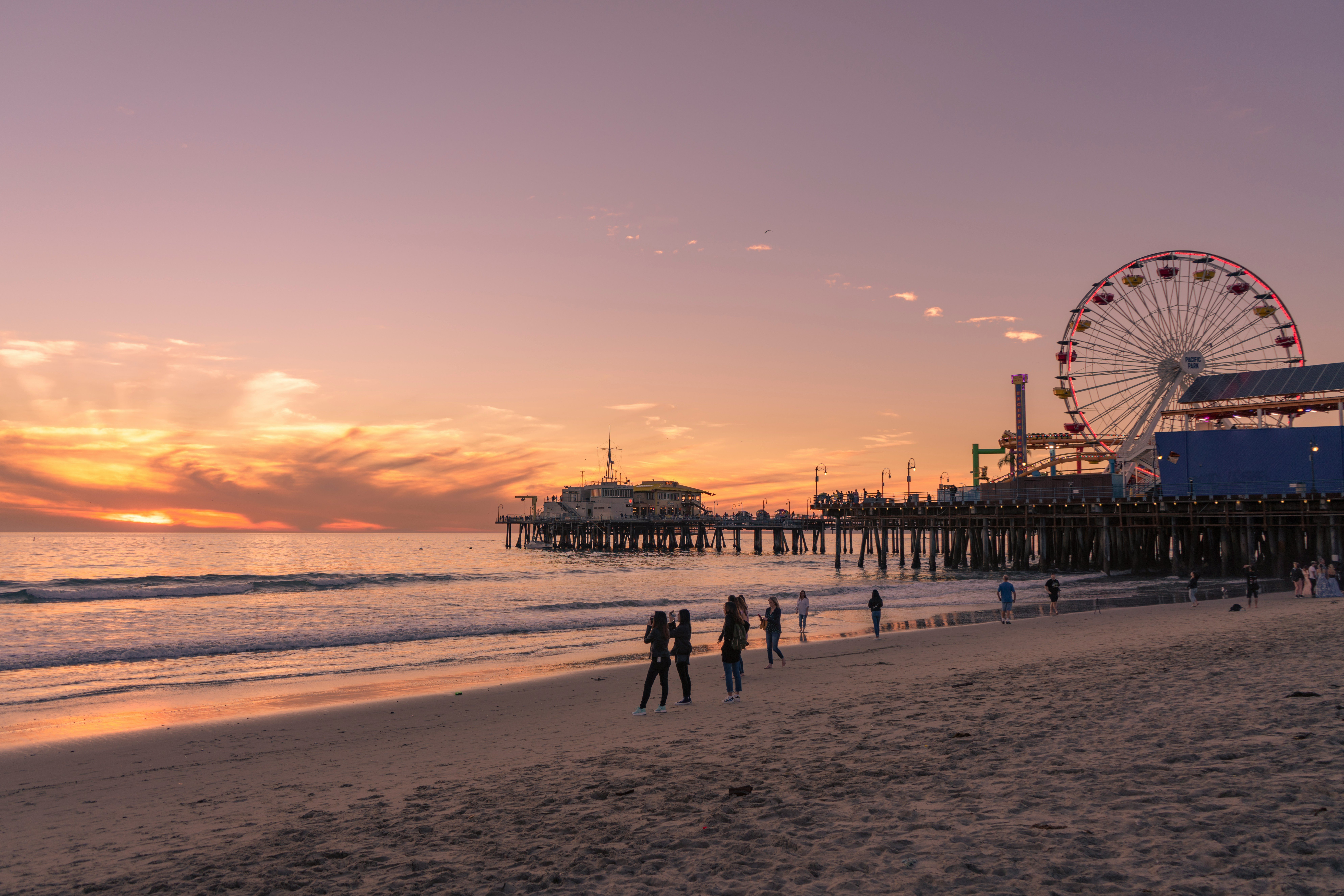 Sunset over the ocean with a lit Ferris wheel on a pier to the right and people walking along the shore.