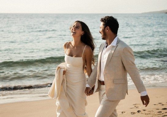 Couple walking hand in hand on the sandy beach in light beige outfits, smiling as waves roll in behind them at sunset
