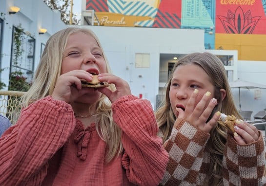 Two girls eating cookies outdoors in front of a bright, colorful mural on a building.